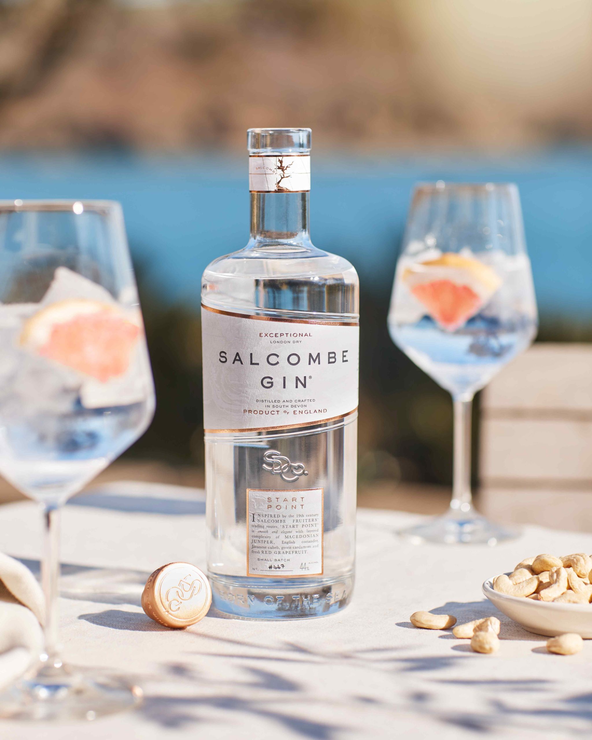 Bottle of Salcombe Gin with two gin and tonics and the sea in the background as the sun shines down as part of a gin advertising campaign
