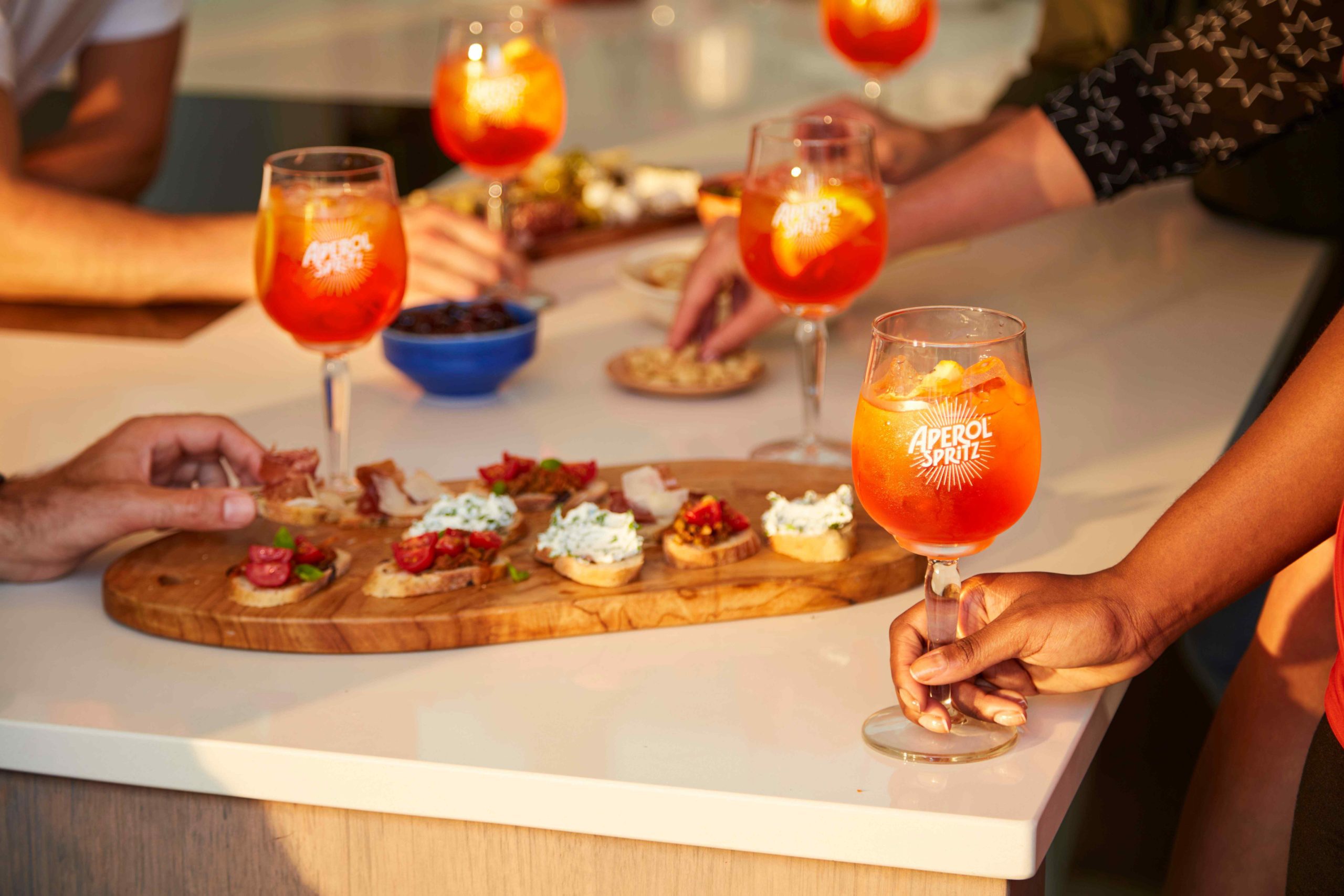 Out Of Home billboard advert by YesMore spirits Marketing Agency for Aperol Spritz showing a hands of a group of friends holding Aperol Spritz with aperitivo dishes around a kitchen counter
