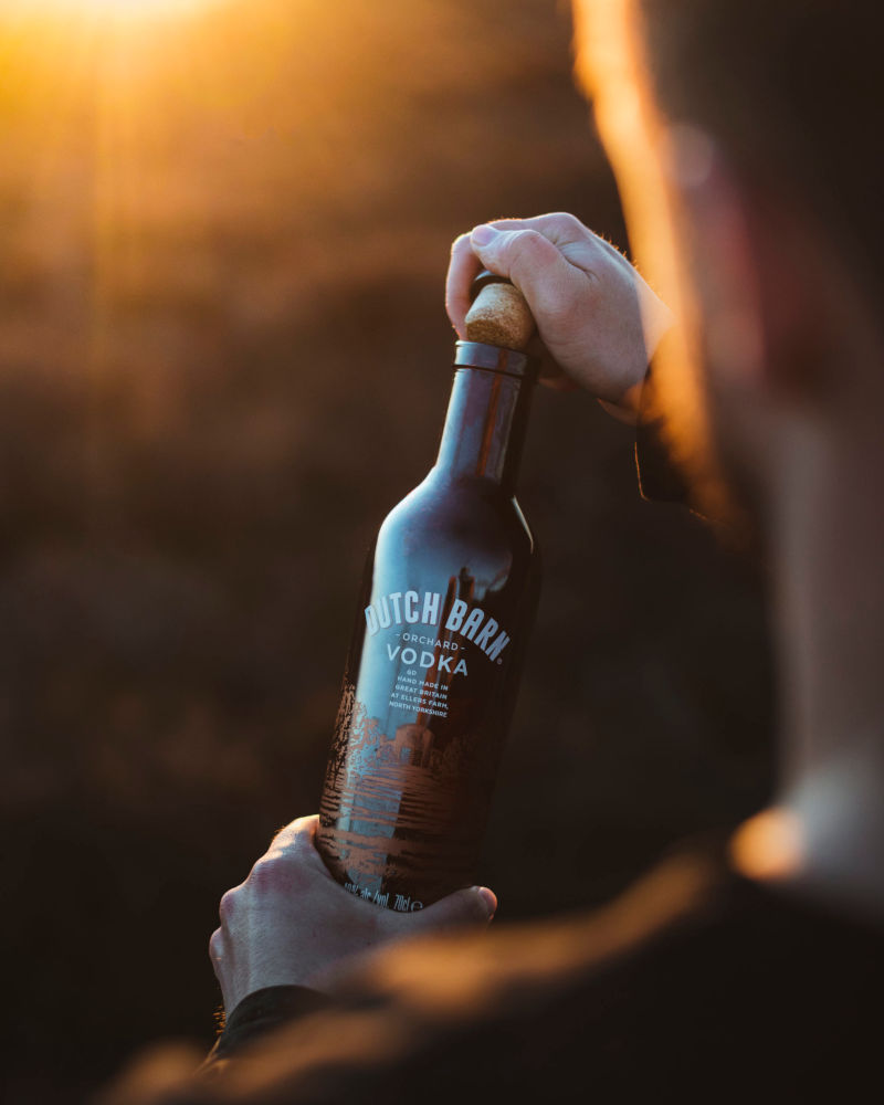 Brown bottle of Dutch Barn Orchard Vodka over the shoulder of someone opening it at sunset, by YesMore Vodka Advertising Agency